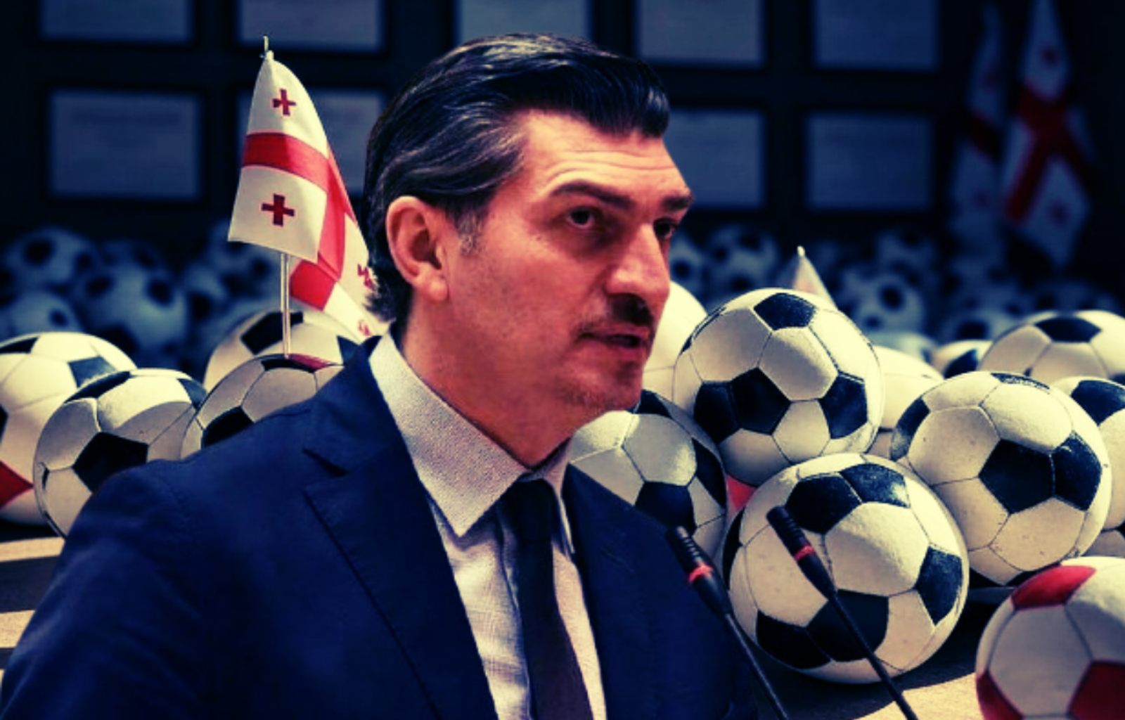 Un uomo in abito scuro, che parla davanti a due microfoni, circondato da palloni da calcio e bandiere georgiane. L'immagine simbolizza il legame tra il passato sportivo del presidente Kavelashvili e la sua controversa elezione politica in Georgia